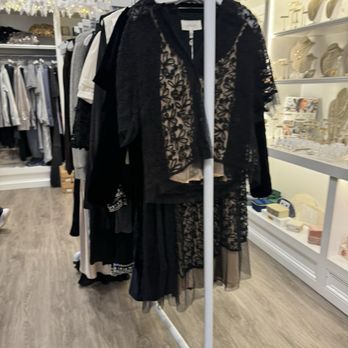 Valentina - A black lace dress with floral embroidery is hanging on a white clothing rack inside a retail store. The dress features sheer lace fabric with intricate floral patterns and is layered over a nude or beige lining. The store has shelves with decorative items and jewelry, and other clothing items are visible on racks in the background. The flooring is wood, and the store is decorated with holiday or festive garlands. - Women's Clothing Near Me - Glencoe, Illinois