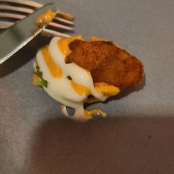 Para Maria - Patatas Bravas - A close-up image shows a fried food item, likely a potato wedge or similar, placed on a gray plate. The item is topped with a drizzle of white sauce and yellow sauce, possibly mayonnaise and mustard or cheese. A fork with some sauce residue is positioned above the food, indicating it is ready to be eaten. The background includes a blurred surface, suggesting a casual dining setting. - Wine Bars Near Me - Boston, Massachusetts