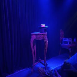 The image shows a small performance stage with dark curtains as the backdrop. On the stage, there is an ornate carved wooden table and a matching chair, both with intricate detailing. A small box and a bottle are placed on the chair, and a hat with a card on top is positioned on the table. The stage is lit with blue lighting, creating a moody atmosphere. In the foreground, a person is seated with their legs crossed, wearing sneakers and holding a drink, suggesting an audience member at a magic or comedy show.