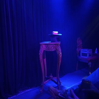 Trickery - The image shows a small performance stage with dark curtains as the backdrop. On the stage, there is an ornate carved wooden table and a matching chair, both with intricate detailing. A small box and a bottle are placed on the chair, and a hat with a card on top is positioned on the table. The stage is lit with blue lighting, creating a moody atmosphere. In the foreground, a person is seated with their legs crossed, wearing sneakers and holding a drink, suggesting an audience member at a magic or comedy show. - Performing Arts Near Me - Chicago, Illinois