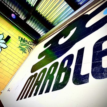 Marble Brewery - The image shows an interior wall of Marble Brewery featuring a large, stylized logo with the word 'MARBLE' painted in bold, black letters on a white background. The wall is part of the brewery's interior decor, with graffiti art and colorful lighting visible in the background, including a yellow brick wall with additional graffiti and artwork. The ceiling has metallic panels with multicolored lighting effects, creating a vibrant atmosphere. - Breweries Near Me - Albuquerque, New Mexico