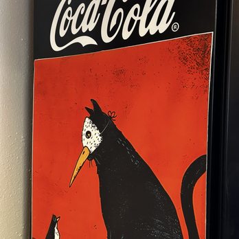 Blackbird Coffee House - A Coca-Cola poster displays a black cat with a white face and a long orange beak, looking at a small bird on a red background. - Coffee & Tea Near Me - Albuquerque, New Mexico