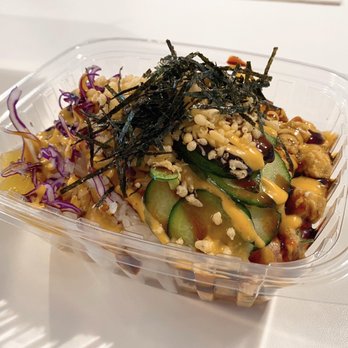Big Poké - Aloha Bowl - A poke bowl in a clear plastic container contains sliced cucumbers, shredded purple cabbage, shredded seaweed, and toppings such as crispy rice, sauces, and possibly fried chicken pieces. The ingredients are arranged with visible sauces drizzled over the top, and the bowl is set against a plain background. - Poke Near Me - Lowell, Massachusetts