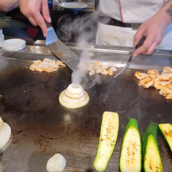 Benihana - Volcano steam!!! - A chef prepares food on a teppanyaki grill, cooking shrimp, onion, and zucchini with steam visible. - Sushi Bars Near Me - Concord, California