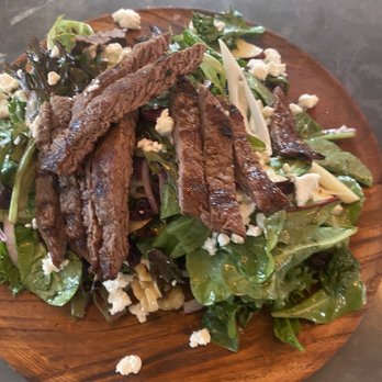 Tiesto's Restaurant - A salad with grilled meat slices, mixed leafy greens, crumbled cheese, and sliced vegetables is presented on a round wooden plate. The greens include various types of lettuce and herbs, with some cheese crumbles scattered throughout. The grilled meat appears cooked and sliced, placed on top of the salad. The presentation emphasizes fresh ingredients and a rustic serving style. - Latin American Near Me - Patchogue, New York