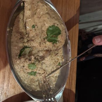 Poppi's Anatolia - Chicken Korma - A serving of chicken korma with a thick, creamy sauce containing herbs and spices is presented in a metal boat-shaped dish. The dish is garnished with cilantro leaves and is placed on a wooden table, with a fork inserted into the food. The presentation suggests a flavorful Indian cuisine dish, likely from a restaurant setting. - Indian Near Me - Eugene, Oregon