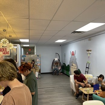 Starry Moon - Inside - The image shows the interior of a bubble tea shop with several customers. Some are standing at the counter, while others are seated on chairs and couches. The decor includes wall art of a large cartoon character and playful elements like plushies and a cotton candy machine. The ceiling has decorative hanging lights, and the space appears bright and inviting, with a mix of seating options and a casual atmosphere. - Bubble Tea Near Me - Albuquerque, New Mexico