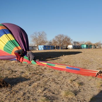 Fly Albuquerque - Fly Albuquerque  flyalbuquerque.com - Two individuals are handling a deflated, multicolored hot air balloon on a flat, open field with a clear blue sky. - Hot Air Balloons Near Me - Albuquerque, New Mexico