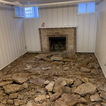 Gumer Concrete LLC - A basement with white paneled walls, small windows, and a brick fireplace, with the floor dug up exposing dirt and rocks. - Masonry/Concrete Near Me - Denver, Colorado