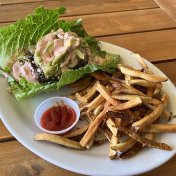 Project Burger - Special Burger but lettuce wrapped! Bomb! - A white oval plate holds a portion of French fries, a lettuce-wrapped burger topped with avocado and sauce, and a small cup of ketchup. The plate is placed on a wooden table, and the burger includes fresh green lettuce and a beef patty. The fries are golden-brown and slightly crispy. The setting appears casual, likely in a restaurant or cafe. - American Near Me - Alameda, California