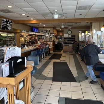 Cafe Olympic - View from front door. - The image shows the interior of a casual dining restaurant with a tiled floor and a ceiling with ceiling fans and recessed lighting. Customers are seated at the counter and in booths along the windows. The counter area has a menu board and a television mounted on the wall. The kitchen and service area are visible in the background, with staff working behind the counter. The space includes wooden accents and a clock on the wall. - New American Near Me - Crystal Lake, Illinois