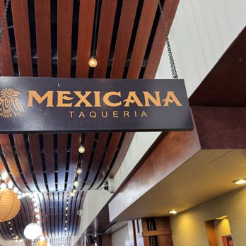 Mexicana - A rectangular sign with the text 'Mexicana Taqueria' hangs from the ceiling, featuring a stylized bird logo, inside a restaurant with wooden ceiling panels and string lights. - Mexican Near Me - Berkeley, California