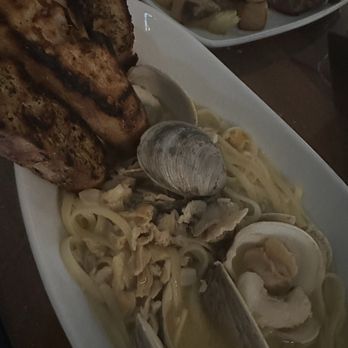 Sogno Restaurant - Linguini and clams - A white rectangular plate holds linguini pasta with clams in a light sauce, garnished with shellfish. Two grilled fish fillets are placed alongside the pasta. In the background, another plate with potatoes and a meat dish is partially visible. The setting appears to be a restaurant with a dark table surface. - Wine Bars Near Me - St. Augustine, Florida