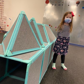 Over the Moon - Building with large magnetic tiles - A young girl with long hair, wearing a face mask, striped long-sleeve shirt, floral skirt, black leggings, and colorful socks, stands next to a large pyramid-shaped structure made of magnetic tiles with gray felt panels and turquoise edges. The indoor play area features a metallic climbing wall with colorful holds and a neutral-colored wall in the background. - Indoor Playcenter Near Me - Cary, North Carolina