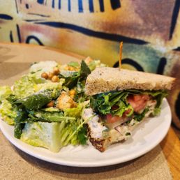 Caesar Salad - A plate features a half sandwich with visible lettuce, tomato, and cheese on whole grain bread, secured with a toothpick. Next to it is a side salad with mixed greens, croutons, and small round toppings, possibly pearls or seeds. The background includes a colorful, abstract wall design, and the plate is placed on a wooden surface.