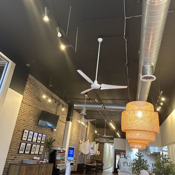 Rumi Middle Eastern Grill - The restaurant interior features a black ceiling with exposed metallic ductwork and multiple ceiling fans. There are track lights and small spotlights illuminating the space. A large woven pendant light hangs prominently, providing ambient lighting. The walls include a brick accent wall with framed pictures and a mounted TV. The seating area has tables and chairs, with some patrons visible. The floor is wooden, and there are decorative plants near the entrance. The overall design combines industrial and modern elements. - Mediterranean Near Me - Chicago, Illinois