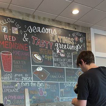 First Watch - Colorful menu options - A man with dark hair and a black shirt is looking at a large chalkboard menu with colorful writing inside a cafe or restaurant. - Cafes Near Me - Tucson, Arizona