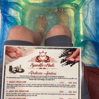Sparkle Nails - The image shows a person with their feet submerged in a foot spa at Sparkle Nails, a nail salon. The foot spa is lined with a blue plastic cover, and the person's knees are visible. A pedicure menu is placed in front of them, listing different pedicure services, including basic, mini, deluxe, and collagen pedicures, with prices ranging from $30 to $80. The menu details the benefits of each treatment, and the salon's focus on pedicure services is evident. The setting appears clean and organized, with a towel and a faucet visible at the top of the image. - Nail Salons Near Me - Ceres, California