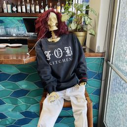 Brunch - Skeleton mannequin with red hair, wearing a black sweatshirt with 'F.O.B KITCHEN' printed on it, seated on a wooden chair in a bar or restaurant interior. The background includes a bar counter with bottles, glasses, and a potted plant near a window. The setting features teal and blue hexagonal tiles on the wall and a window with patterned glass.