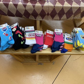Prisco's - The image shows several stacks of socks with various designs, including cartoon characters like SpongeBob SquarePants, and brand logos such as Pepsi and Coca-Cola. The socks are arranged in neat piles on a wooden table, with some featuring colorful patterns and others with character-themed designs. The setting appears to be indoors, possibly in a store or market, with a table covered by a patterned cloth visible at the top of the image. - Delis Near Me - Bridgewater, Massachusetts