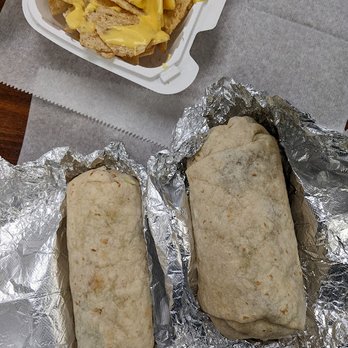 Tacocat - "Chicky Creama" and "Tacocat Carne Asada". - Two burritos wrapped in aluminum foil are placed on a surface, with a side of nachos topped with melted cheese in a takeout container. - Mexican Near Me - Springville, New York