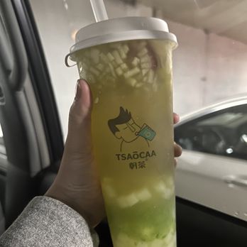 Tsaocaa - Japanese Melon Green Tea W. Nata Jelly - A person holds a large, clear plastic cup filled with green tea and diced fruit, with a white lid and straw. The cup features a cartoon logo of a person drinking with the text 'TSAOCAA' and Chinese characters. The background shows part of a vehicle interior and a parking lot outside, indicating the drink is being enjoyed in a car or near a parking area. - Bubble Tea Near Me - Pittsburgh, Pennsylvania