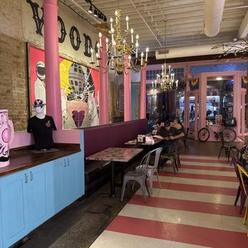 Voodoo Doughnut - The image shows the interior of a cafe or restaurant with a mix of vintage and modern decor. Pink columns support the ceiling, which has exposed pipes and beams. Chandeliers hang from the ceiling, providing lighting. A mural with colorful artwork is visible on the wall behind the seating area. Customers are seated at tables, some reading or using devices. The floor has a red and white striped pattern, and there are bicycles near the entrance. The space has a cozy, eclectic atmosphere. - Donuts Near Me - Austin, Texas