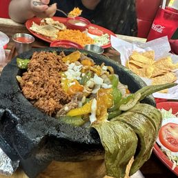 Comida molcajete - A large black stone molcajete contains seasoned ground meat, sautéed bell peppers, onions, and melted cheese, placed on a wooden serving board. Surrounding the molcajete are side dishes including rice, tortillas, and chips. In the background, a woman is eating from a red plate with rice, tortillas, and condiments, with a Coca-Cola cup nearby. The setting appears to be a casual Mexican restaurant with vibrant decor.