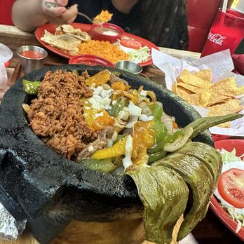El Nopal - Comida molcajete - A large black stone molcajete contains seasoned ground meat, sautéed bell peppers, onions, and melted cheese, placed on a wooden serving board. Surrounding the molcajete are side dishes including rice, tortillas, and chips. In the background, a woman is eating from a red plate with rice, tortillas, and condiments, with a Coca-Cola cup nearby. The setting appears to be a casual Mexican restaurant with vibrant decor. - Mexican Near Me - Dover, Delaware