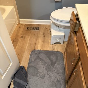 Williamson Management - A bathroom features a white toilet with a closed lid, positioned against a blue wall. To the right of the toilet is a wooden cabinet with a white countertop. A gray bath mat is placed on the wood floor in front of the toilet and cabinet. A vent is visible on the floor near the door, and a towel is on the floor near the door. The bathroom has a partial view of a bathtub with beige tiles on the left side. - Property Management Near Me - Bensenville, Illinois