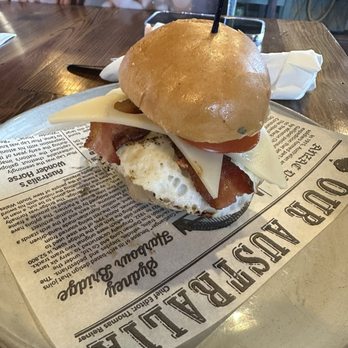 Byte & Coffee - A breakfast sandwich featuring bacon, fried egg, cheese, and tomato slices is placed on a plate lined with newspaper-style paper. The sandwich has a toasted bun and is set on a wooden table in a restaurant setting. The background includes a smartphone and napkins, indicating a casual dining environment. - Coffee & Tea Near Me - Pompano Beach, Florida