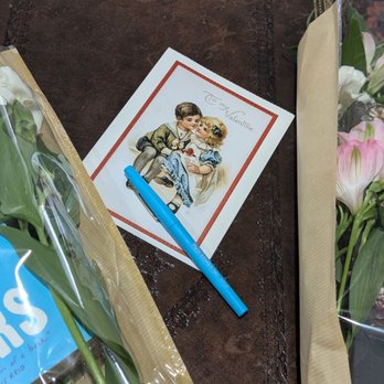 The Noble Capybara - Valentine event "Books and Blooms" - A Valentine's Day card with an illustration of a boy and girl kissing is positioned on a dark wooden table. The card has the message 'To my Valentine' written on it. Around the card, there are bouquets of white and pink flowers wrapped in paper and plastic, along with a blue book and a pen placed on the card. - Used Bookstore Near Me - Lisle, Illinois