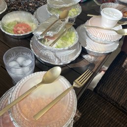 Multiple disposable aluminum and plastic containers with salads, including lettuce and tomato, are placed on a dark wooden dining table. Gold-colored plastic utensils, including spoons and forks, rest on the containers. A glass of ice water with ice cubes is also visible. The background features various condiments, bottles, and small items, indicating a casual dining or takeout setting.