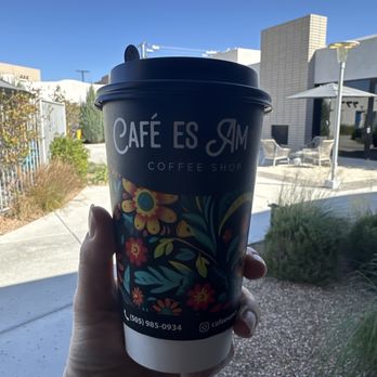 Cafe es Amor - Super cute to go cup with my amorcito latte inside! - A person holds a coffee cup with a floral design outside a coffee shop on a clear day, with outdoor seating and modern buildings in the background. - Coffee & Tea Near Me - Albuquerque, New Mexico