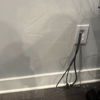 FixTman - Washington - Poor paint job, paint on floor and outlet - The image depicts a wall with noticeable damage, including peeling paint and cracks. An electrical outlet is mounted on the wall, with two black cords hanging from it and resting on the floor. The floor is dark-colored, and there is a baseboard along the bottom of the wall. The scene suggests a need for repair or maintenance. - Furniture Assembly Near Me