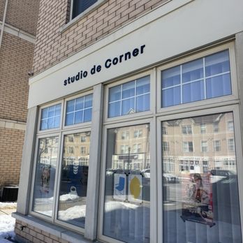 Studio De Corner - The image shows the exterior of Studio De Corner, a business located in Markham, ON. The storefront has large windows with reflections of nearby buildings and parked cars. The signage above the windows displays the business name in lowercase letters. The building is constructed with brick and concrete, and there is snow on the ground indicating winter conditions. - Cafes Near Me - Markham, Ontario