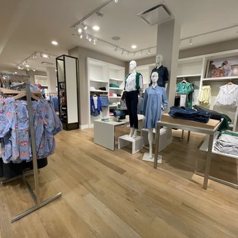 LOFT - The image shows a retail clothing store interior with mannequins dressed in casual outfits, including shirts and pants. The store has wooden flooring, white shelving units, and clothing racks with various garments. The mannequins are arranged near the center, with shelves of folded clothes and framed photographs on the walls. The lighting is bright, illuminating the display area. - Accessories Near Me - Tampa, Florida