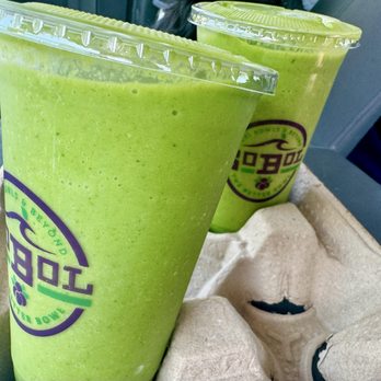 SoBoL - Two green smoothies in clear plastic cups with lids are positioned on a tray, possibly in a restaurant or cafe. The smoothies are vibrant green and appear thick and blended. The cups have a logo with purple and green colors. The background includes a gray surface and part of a seat or table, indicating an indoor or vehicle setting. - Juice Bars & Smoothies Near Me - Westhampton Beach, New York