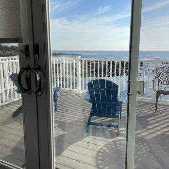 Perkins Cove Cottage - The image shows a balcony with a white railing, overlooking a rocky shoreline and the ocean under a partly cloudy sky. There are two blue chairs and a white chair on the balcony, with shadows cast by the railing. The photo is taken from inside, through a glass door with handles and a lock visible. The scene suggests a coastal setting, possibly part of a seaside property or vacation rental. - Vacation Rentals Near Me - Ogunquit, Maine