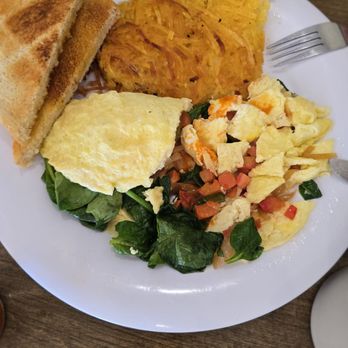 Baja Cafe - Build your own omelette - A breakfast plate includes two slices of toasted bread, a portion of hash browns, scrambled eggs, and a vegetable salad with leafy greens, chopped tomatoes, and pieces of scrambled eggs. The food is arranged on a white round plate placed on a wooden table, with a fork visible on the right side of the plate. - Breakfast & Brunch Near Me - Tucson, Arizona