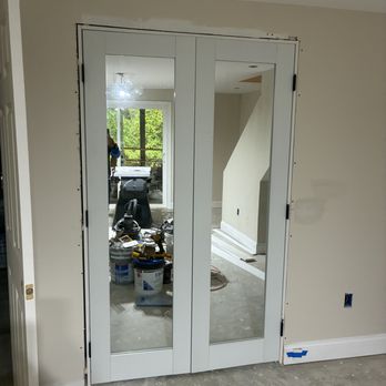 Infinite contracting - Finished basement- Drywall, Paint, Trim,Doors - The image shows a pair of double glass doors installed in an interior wall of a home under renovation. The doors have white frames and are partially open, revealing a room with construction supplies, tools, and a person working outside through a window. The surrounding wall has visible gaps and unfinished edges, indicating ongoing work. The floor is unfinished concrete, and the wall has some electrical outlets and wiring preparations. - Painters Near Me - Forest Hill, Maryland