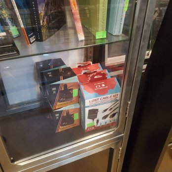 Killen Enterprises - The image depicts a glass display case with metal framing, containing gaming accessories such as a 'Lost Cable Kit' and boxed items labeled 'Trooper.' The case also holds books on topics like comics and collectibles, with some green price tags visible. The setting appears to be a retail store specializing in games, comics, and related products, operating Monday through Saturday. - Toy Stores Near Me - Springfield, Illinois