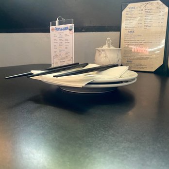 Ebisu - Clean table wow - A white plate with black chopsticks and napkins is positioned on a dark, smooth restaurant table. In the background, there are menus standing upright and a small bowl, indicating a dining setting. The image captures a clean, minimal table setup in a Japanese or Asian-style restaurant. - Sushi Bars Near Me - Providence, Rhode Island