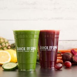 Raw Juice Mix - Two juice cups, one green and one red, are placed on a surface with fresh fruits and vegetables nearby, including lemon, cucumber, pineapple, carrots, and beets.