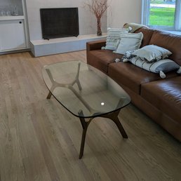 floors! - A living room features a glass coffee table, a brown leather sofa with decorative pillows, and a window with a view of trees and grass.
