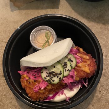 Take a Bao Eats - Crispy Spicy Aioli Chicken Bao - A bao sandwich features fried chicken topped with sliced cucumbers sprinkled with black sesame seeds, pickled vegetables, and a steamed bun, all arranged in a black takeout container. A small cup of sauce is included, and the background shows a brown paper bag and other containers on a textured surface. - Asian Fusion Near Me - Chicago, Illinois