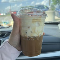 crème Cream Brulee Coffee - A person holds a plastic cup of iced coffee from Sip Munch Cafe, with a clear lid, inside a vehicle. The background shows a parking lot and blue sky.