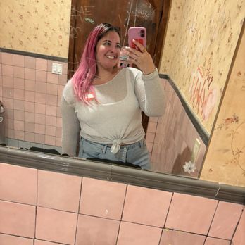 Fox’s Lounge - Bathroom Selfie - A woman with shoulder-length pink hair, wearing a white long-sleeve top tied at the waist, is smiling and holding a smartphone with a pink case, taking a selfie in a bathroom mirror. The bathroom has pink tiled walls, graffiti on the door behind her, and a floral wallpaper on the upper part of the wall. The mirror reflects her upper body and part of the bathroom interior. - Dive Bars Near Me - Miami, Florida