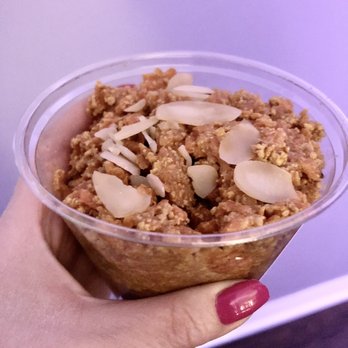 Khokha Eatery - Carrot Halwa - A transparent plastic cup filled with carrot halwa, a traditional Indian dessert made from grated carrots, sugar, and ghee, is shown. The dessert is topped with sliced almonds, adding texture and flavor. The hand holding the cup has red nail polish, and the background is a plain, light-colored surface. - Pakistani Near Me - Ottawa, Ontario