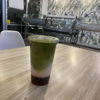 Rewind - Strawberry Matcha Latte - A layered matcha drink features distinct green, white, and red layers in a clear plastic cup with a lid. The drink is on a light-colored wooden table inside a cafe, with a background of patterned wallpaper, lighting fixtures, and a glass display case. - Bubble Tea Near Me - Chandler, Arizona